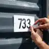 Handen die witte huisnummer sticker op donkere metalen zeecontainer aanbrengen met gereedschap in daglicht
