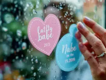 Waterbestendige baby-aankondigingsstickers worden op regenachtig raam geplakt, hand met trouwring drukt aan