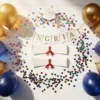Kleurrijke afstudeerdecoraties met gouden en blauwe ballonnen, confetti en diploma-uitsneden symmetrisch gerangschikt op witte houten tafel