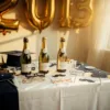 Elegante afstudeertafel met gouden folieballonnen, gepersonaliseerde champagneflessen en fotobooth-accessoires in warm zonlicht
