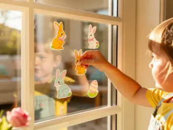 Kind plaatst kleurrijke paashaas en ei stickers op woonkamerraam met ochtendlicht en zachte schaduwen