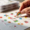 Kleurrijke feestelijke stickers netjes gerangschikt op wit perkamentpapier met vingers die een regenboog verjaardagssticker optillen