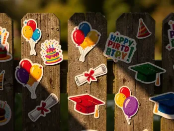 Kleurrijke verjaardag- en afstudeerstickers op verweerde houten schutting met regendruppels, heldere kleuren in ochtendlicht